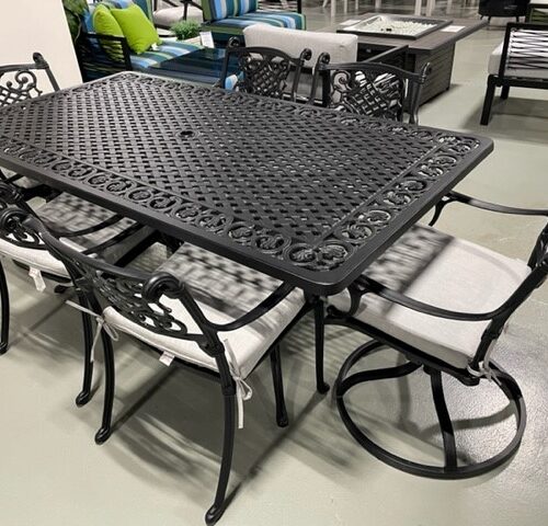 A Gensun Michigan black metal patio dining set with a latticed design. The set includes a rectangular table and four chairs with white cushions, displayed in an indoor showroom.