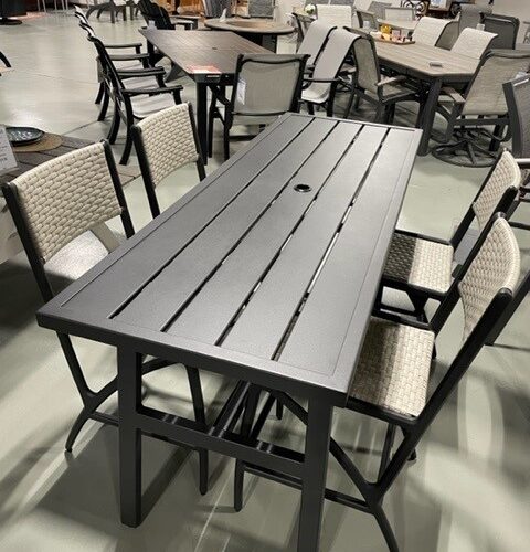 A showroom displaying a modern Gensun Amari outdoor dining set with a rectangular grey slatted table and surrounding wicker chairs with gray cushions, set on a concrete floor.