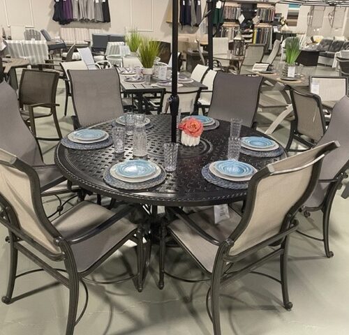 A Gensun Bel Air patio dining set display in a store, featuring a round, black table and six matching chairs with gray cushions, elegantly set with plates and glasses, and a small floral