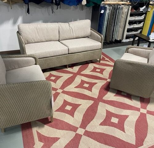 A modern living room setup featuring a beige sectional sofa and two matching armchairs on a red and beige patterned rug, displayed in a showroom with various fabrics in the background.