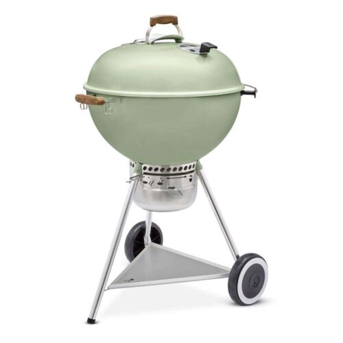 A 70th Anniversary light green Weber Grill Kettle with a lid, featuring wooden side handles and a bottom shelf, is mounted on a tripod stand with wheels for mobility, against a white background.