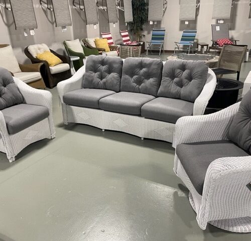 A furniture showroom displaying a gray tufted sofa flanked by two matching armchairs with white wicker frames, placed on a large reflective surface, enhancing the reflections.