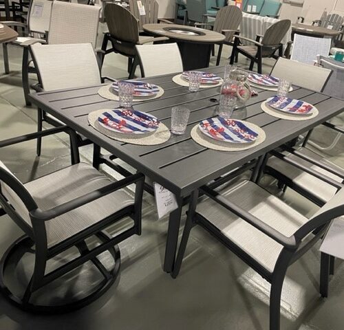 A showroom display of a modern Gensun Echelon outdoor dining set featuring a gray table and white-framed chairs with mesh seating, set with patriotic-themed place settings.
