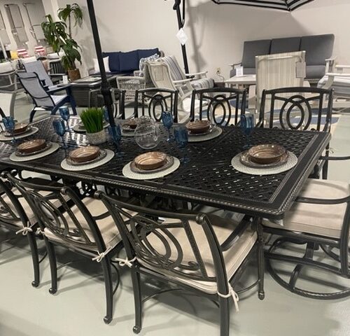 A display of an elegant outdoor dining set at Best Fire Hearth & Patio, featuring a black table and six chairs with cream cushions, set with plates, glasses, and blue decor.