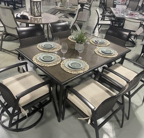 A Gensun Treviso dining set displayed in a store, featuring a square table with a dark top and beige framed chairs on wheels, set with plates, glasses, and a center plant.