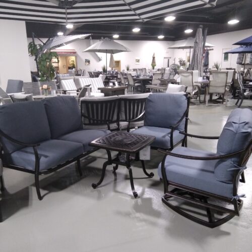 A furniture showroom displays a patio set with blue cushioned chairs and a small table under a large black and white striped umbrella, surrounded by various other furniture pieces.