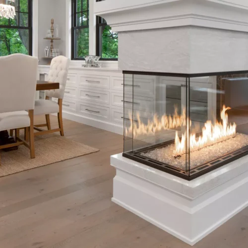 A modern, well-lit dining room with a large table and upholstered chairs features the Enlight Pier Three-Sided Gas Fireplace by Stellar by Heat & Glo prominently in the foreground, adding warmth to the