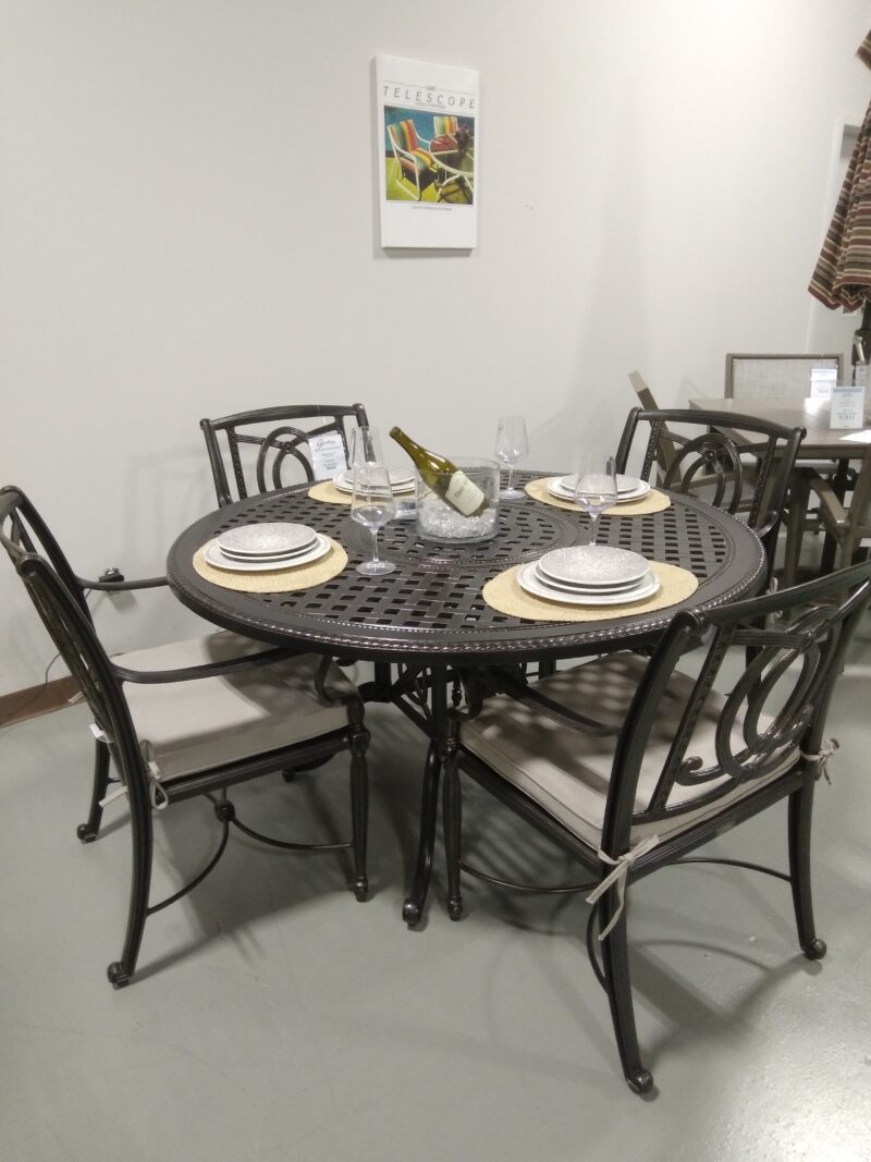 A dining setup featuring a round Gensun Bel Air metal table and four chairs, with place settings for two, including plates and napkins. A wall decor piece hangs above, titled "Telescope