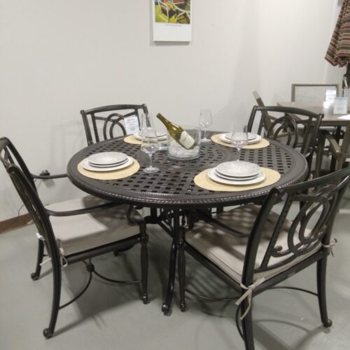 A dining setup featuring a round Gensun Bel Air metal table and four chairs, with place settings for two, including plates and napkins. A wall decor piece hangs above, titled "Telescope