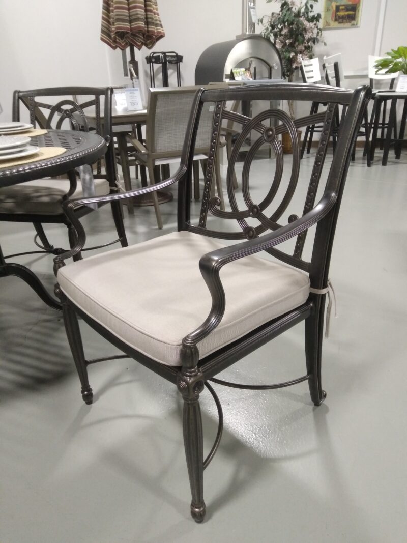 A Gensun Bel Air metal dining chair with ornate backrest and beige cushion, situated on a shiny floor in a room with more furniture in the background.