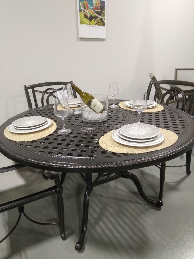A Gensun Bel Air round, black metal dining table set for four, with woven placemats, white plates, and wine glasses. A bottle of wine lies in the center, against a