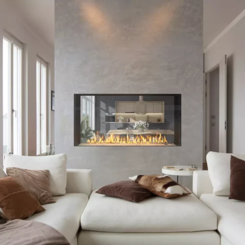 A cozy living room featuring a large white sectional sofa with brown and beige pillows, an Enlight See-Through Gas Fireplace by Stellar by Heat & Glo set in a concrete wall, and soft lighting. There