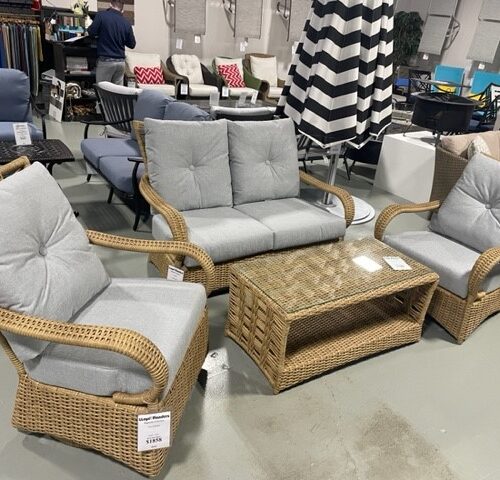 A variety of patio furniture displayed in a store, featuring magnolia wicker chairs with gray cushions, a matching sofa, and a coffee table with a glass top.