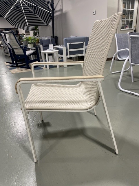 A modern beige Gensun Ventura chair with a woven seat and backrest, and wooden armrests, placed on a gray floor in an indoor setting with blurred furniture and umbrellas in the background