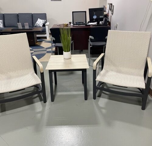 Two Gensun Ventura white outdoor chairs facing each other with a small table between them, atop a light grey floor in a room with soft lighting and furniture in the background.