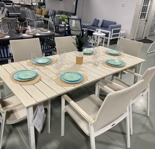 A modern Gensun Ventura outdoor dining set displayed in a showroom, featuring a light wooden table and six white woven chairs, each set with blue plates and clear glasses.