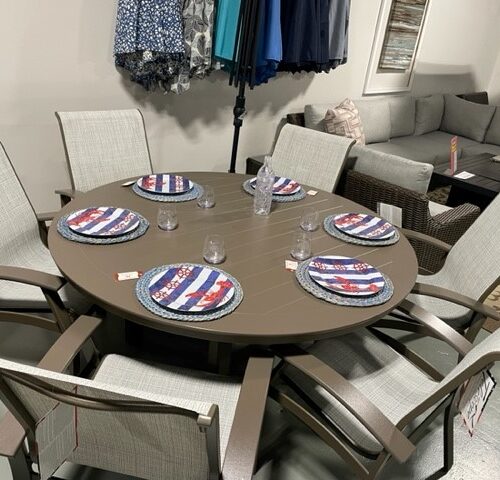 A round dining table set for four with patriotic-themed placemats and clear glasses, surrounded by six Belle Isle Sling chairs, located in an indoor room with clothing racks and a couch in the background