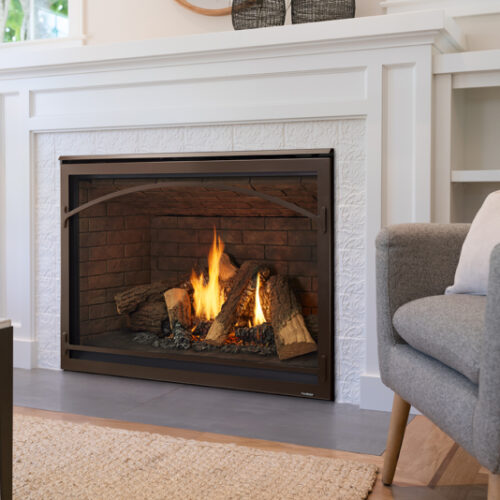 A cozy living room with a Heatilator Caliber Gas Fireplace, featuring realistic logs and flames. A grey sofa is partially visible on the right, with built-in white shelves in the background.
