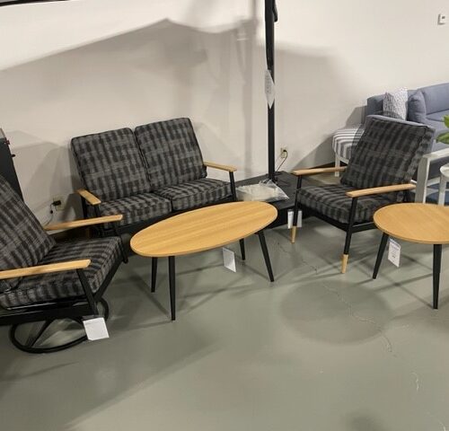 A furniture display featuring two checkered armchairs and a plain armchair from the Welles 4 Person Patio Set by Telescope Casual, centered around three circular wooden coffee tables on a concrete floor.