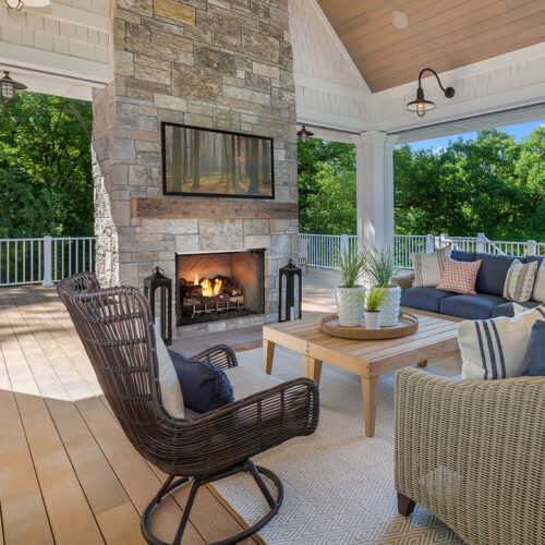 A cozy outdoor patio with a Vesper Outdoor Fireplace, wicker furniture, and wooden floor set against a background of lush green trees under a sunny sky.
