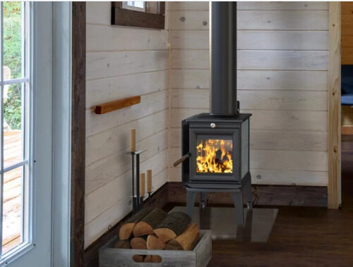 A cozy cabin interior featuring a lit wood stove with visible flames, a neatly stacked wood basket beside it, and light wooden walls that complement a bright outdoor view visible through a window.