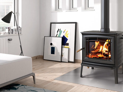 A cozy living room featuring a lit wood-burning stove, a minimalist white couch, and a floor lamp. art supplies and canvases lean against a wall near a large window.